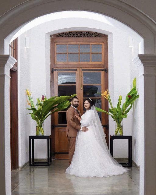 Bride in white wedding gown and groom in tan suit standing in white arched doorway with tropical green plants