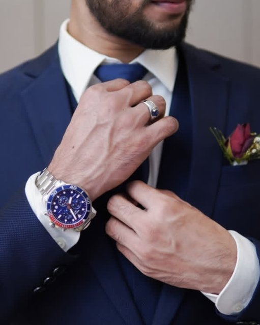 Groom in navy suit adjusting tie with silver watch and red rose boutonnière